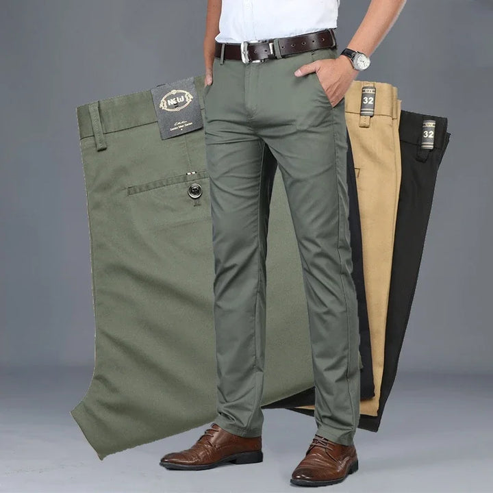 A man wearing olive green dress pants with a tailored fit, brown belt, white shirt, and brown shoes stands in front of three pairs of folded pants in olive green, beige, and black—highlighting their versatile style against a plain grey background.