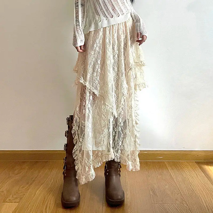 A person stands indoors wearing a white, long-sleeved, distressed sweater and a layered, cream-colored silky lace skirt with an asymmetrical hemline, paired with tall brown boots on a wooden floor.