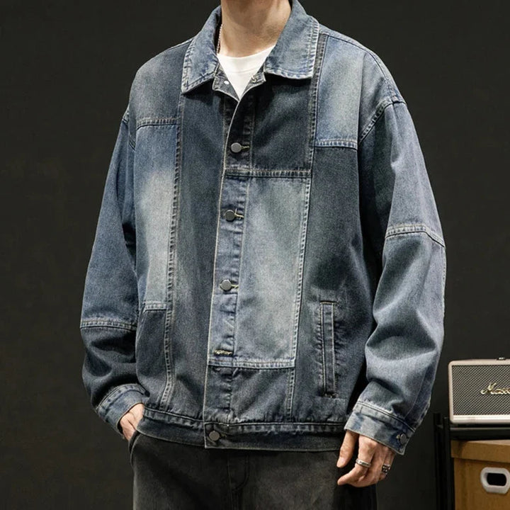 A person wearing an oversized fit, button-up denim jacket with a faded, patchwork design stands with hands in pockets against a dark background. Only the upper body is visible, highlighting the retro style of the denim patchwork.