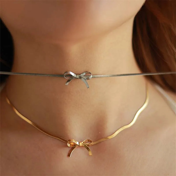 A close-up of a womans neck wearing minimalist jewelry: a silver bow necklace choker and a gold, wavy adjustable pendant with matching bows, both centered at the front.