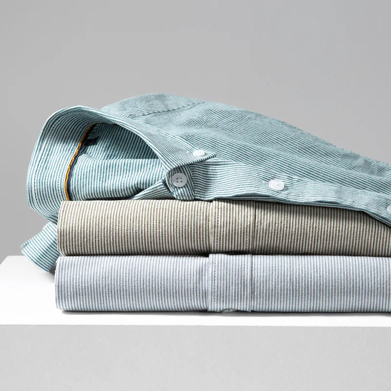 Three neatly folded classic pinstripe shirts in pastel shades (light green, beige, and light gray) are stacked on a white surface against a plain gray background. Each cotton button-up is machine washable for easy care.