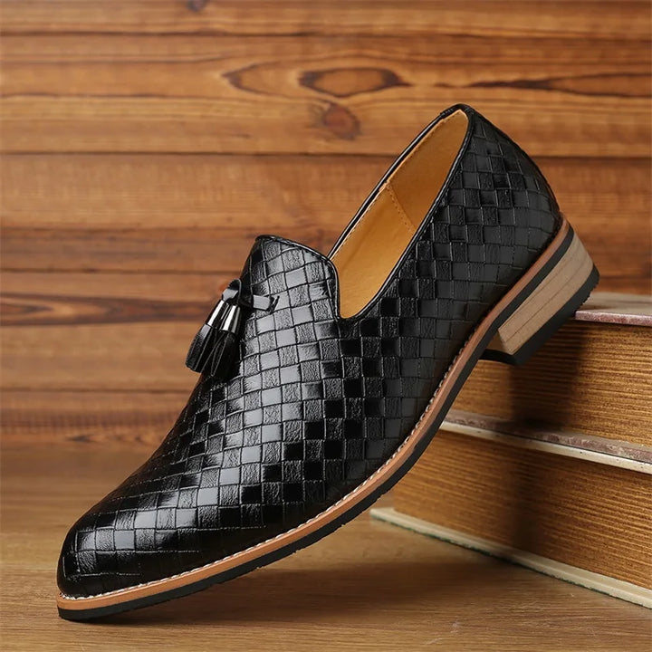A luxury footwear piece, this black real leather shoe features a basketweave pattern with a tassel detail, light brown sole, and tan interior, displayed on wooden steps against a rustic backdrop.
