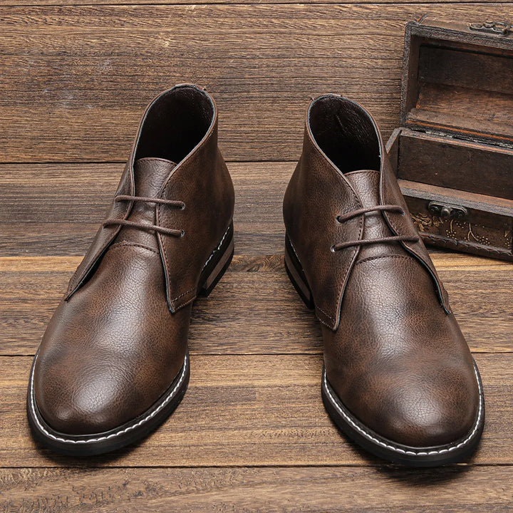 A pair of brown After-Ride Chukka leather boots with black soles and white stitching, placed on a wooden surface next to an open wooden box, showcasing their Western-inspired chukka design.