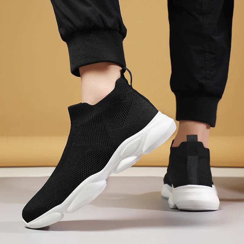 Person wearing black slip-on athletic sneakers with white soles, standing on a gray surface against a tan background. One foot is lifted, highlighting the lightweight, breathable knit fabric and non-slip sole of the flexible shoe.