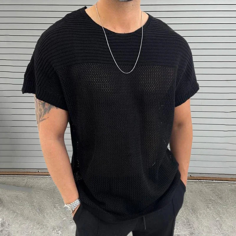 A man wearing a loose black mesh knit tee, black pants, a silver chain necklace, and a watch stands in front of a gray metal panel wall. His hands are in his pockets, and a tattoo is visible on his upper arm.