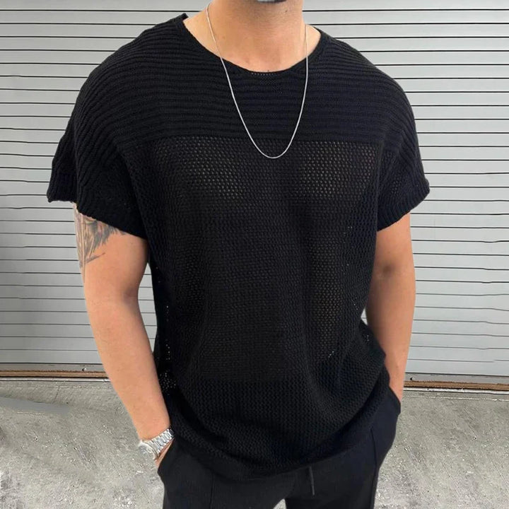 A man wearing a loose black mesh knit tee, black pants, a silver chain necklace, and a watch stands in front of a gray metal panel wall. His hands are in his pockets, and a tattoo is visible on his upper arm.