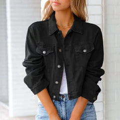 A woman wearing a black, button-up denim jacket over a white top and light blue jeans stands with her hands clasped in front of her, facing slightly to the side. She has long, wavy hair and is accessorized with layered necklaces.