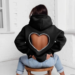 A person sits on a wooden stool facing away, wearing an oversized heart-shaped open back hoodie with rhinestone trim, paired with light blue jeans, in a bright room with white walls.