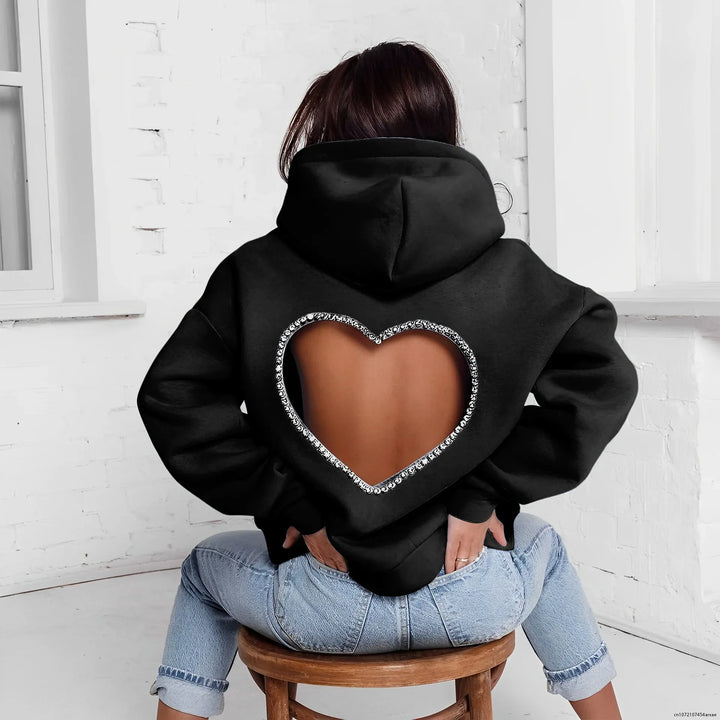 A person sits on a wooden stool facing away, wearing an oversized heart-shaped open back hoodie with rhinestone trim, paired with light blue jeans, in a bright room with white walls.