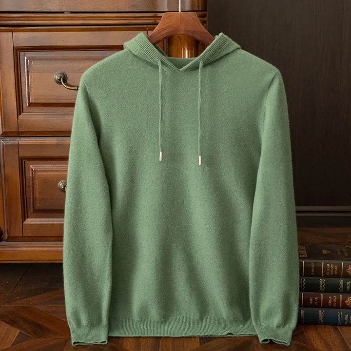 A light green cashmere blend hooded sweater with drawstrings is displayed on a wooden hanger in front of a wooden dresser, with a few books stacked nearby.
