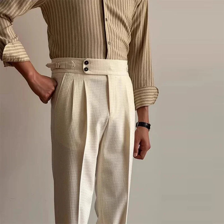 A person wearing a beige striped shirt and high-waisted, luxury tailored pants stands against a plain light-colored background. One hand is in their pocket, and they are accessorized with a dark wristwatch.