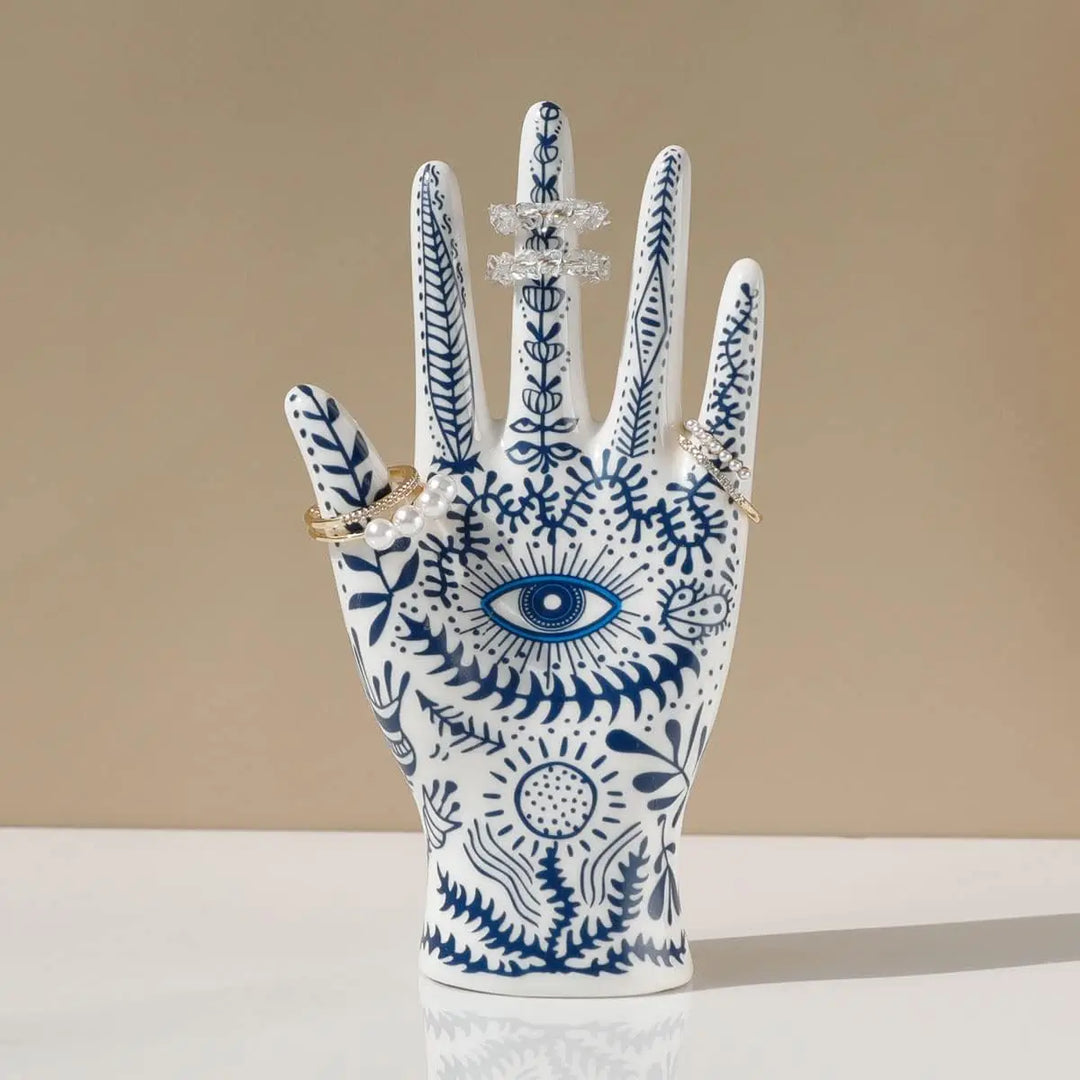 A white ceramic jewelry stand decorated with blue botanical patterns and an eye design displays four sparkly rings on its fingers, set against a neutral beige background—perfect for boho-chic home décor.