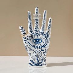 A white ceramic jewelry stand decorated with blue botanical patterns and an eye design displays four sparkly rings on its fingers, set against a neutral beige background—perfect for boho-chic home décor.