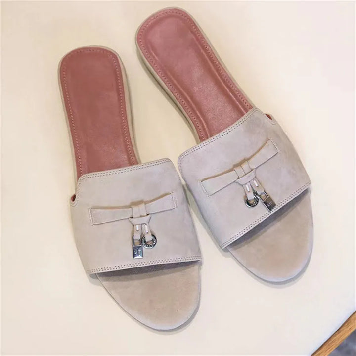 A pair of beige, open-toed slide sandals with a suede finish and pink cushioned insoles, featuring decorative bow accents with small silver charms on the front straps, displayed on a light surface.