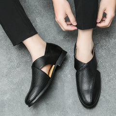 Person wearing modern black genuine leather loafers with a unique cut-out design and durable outsole, sitting on a gray textured floor, adjusting one shoe with their hand. Only the lower legs and hands are visible.