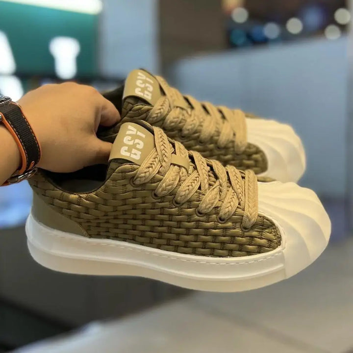 A hand holds a pair of beige woven upper sneakers with thick, white, shell-like toe caps and chunky cushioned soles, displayed indoors with a blurred background.