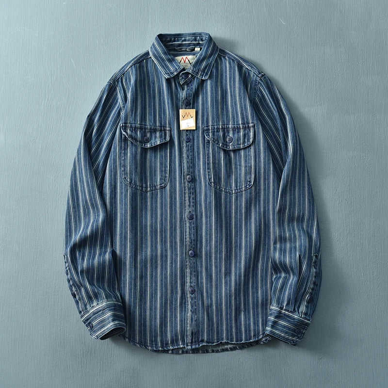 A vintage button-up blue denim shirt with white vertical stripes, two chest pockets with flaps, and a brown tag hanging from the collar, crafted from heavyweight cotton and displayed on a flat light blue surface.