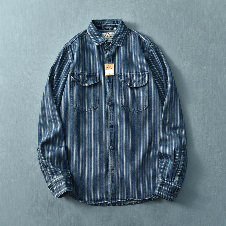 A vintage button-up blue denim shirt with white vertical stripes, two chest pockets with flaps, and a brown tag hanging from the collar, crafted from heavyweight cotton and displayed on a flat light blue surface.