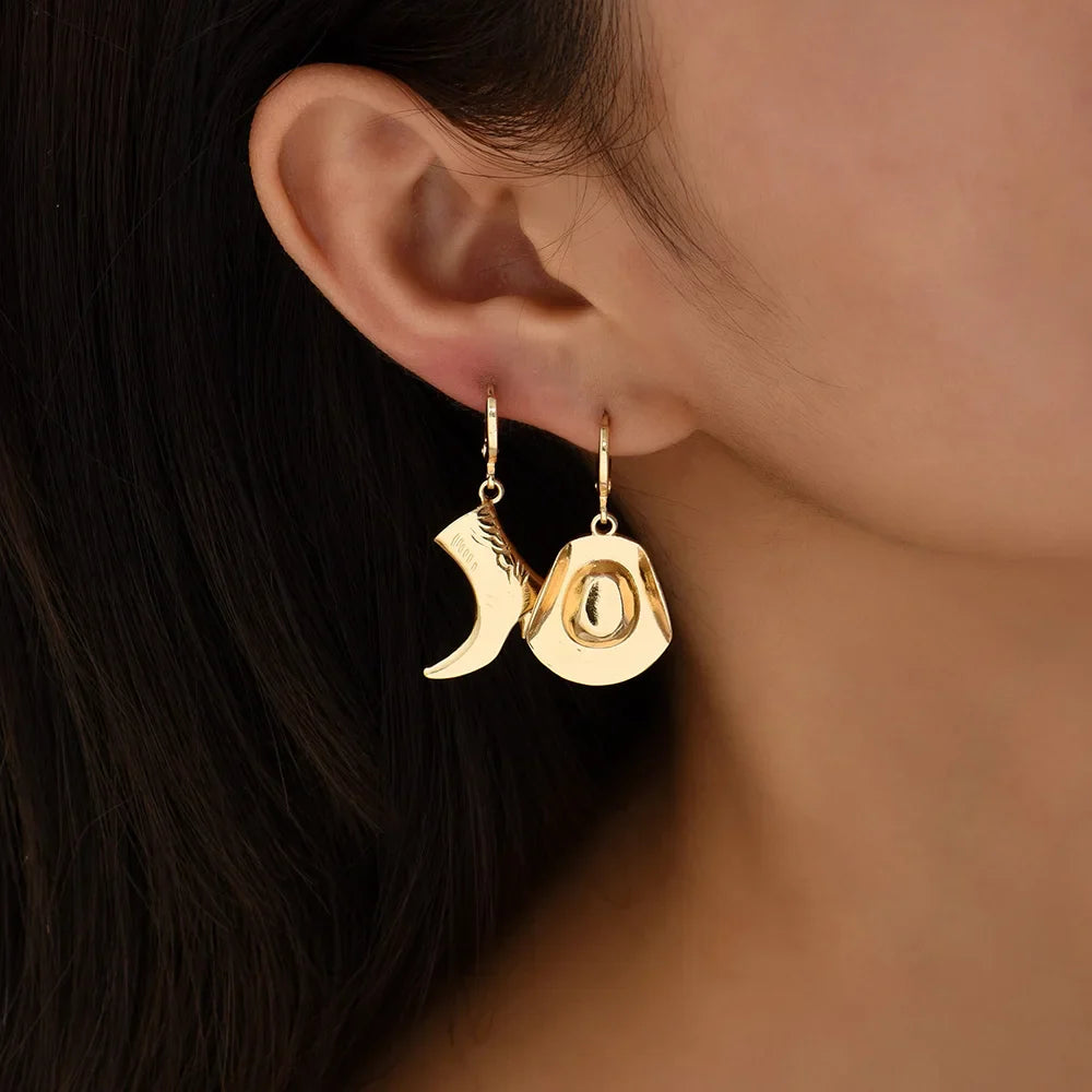 A close-up of a person’s ear wearing mismatched gold cowboy hat earrings and a boot, stylishly showcasing western jewelry against a neutral background.