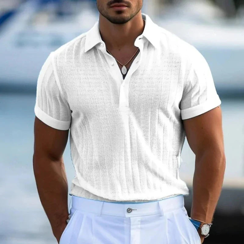 A man wearing a white short-sleeve knit polo shirt crafted from lightweight knit fabric and light blue trousers stands outdoors, with a blurred background suggesting water and boats.