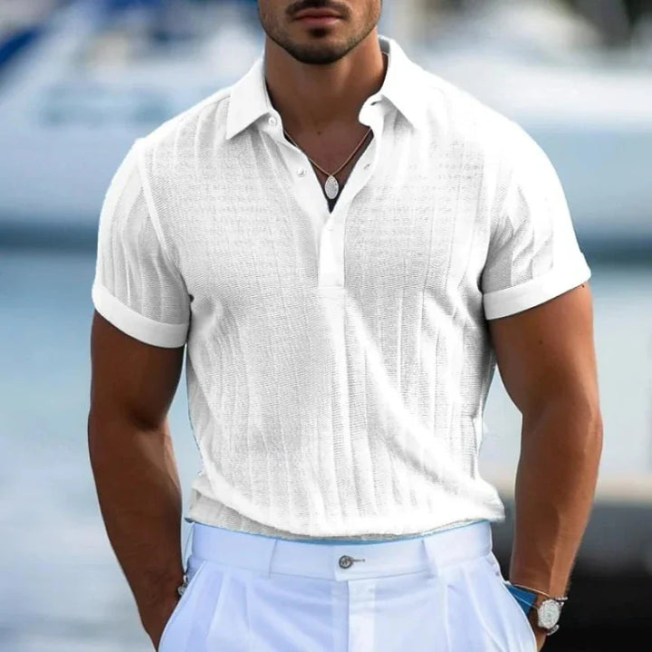 A man wearing a white short-sleeve knit polo shirt crafted from lightweight knit fabric and light blue trousers stands outdoors, with a blurred background suggesting water and boats.