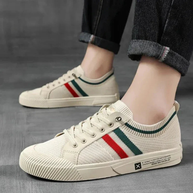 A person wearing cream-colored sneakers with green and red stripes and a durable rubber outsole, paired with rolled-up dark jeans, standing on a gray floor.