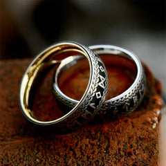 Two silver Viking Rune Rings with intricate engraved designs and gold interiors rest on a textured, reddish-brown brick surface. The couple's amulet rings feature patterns and symbols reminiscent of ancient or fantasy scripts.