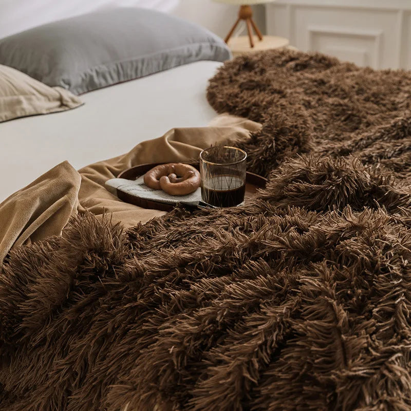 A cozy bed with a warm and cozy Sherpa Blanket, a beige sheet, and a tray holding a glass of dark beverage, a napkin, and a pretzel. Gray and beige pillows are visible in the background.