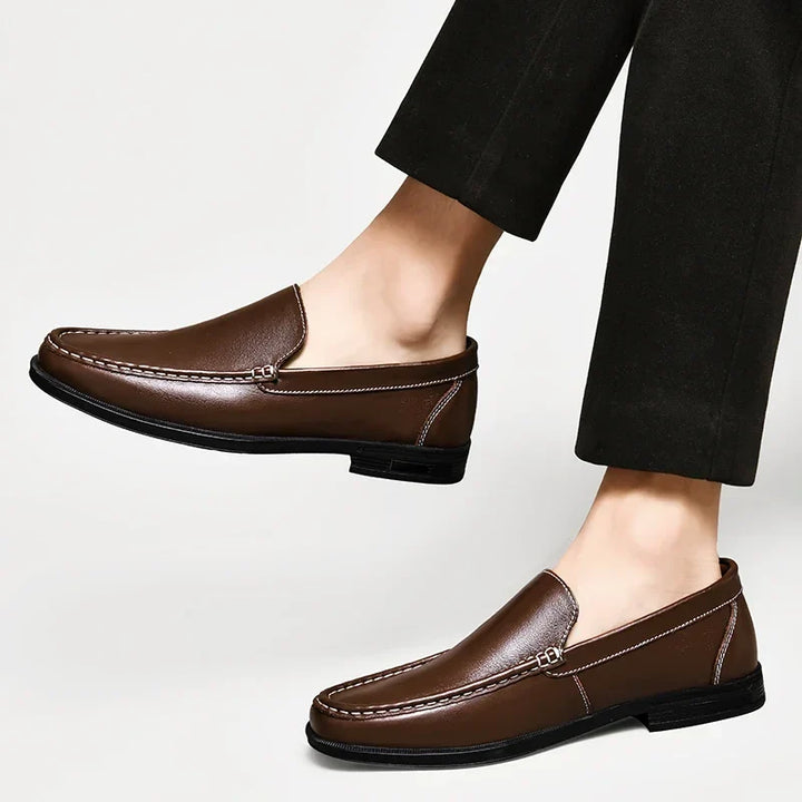 A person wearing dark trousers and classic slip-on brown loafers crafted from genuine leather with visible white stitching, photographed against a plain white background.