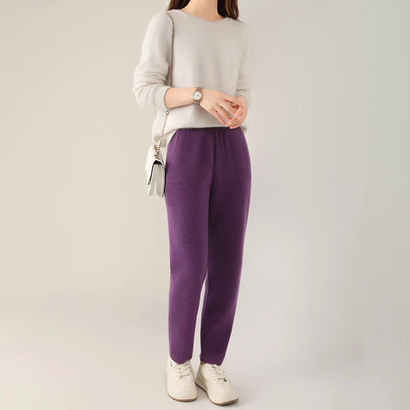 A woman stands against a plain background wearing a light sweater, purple pants with a slim tailored leg, white sneakers, a small shoulder bag, and a wristwatch. Her face is not visible.