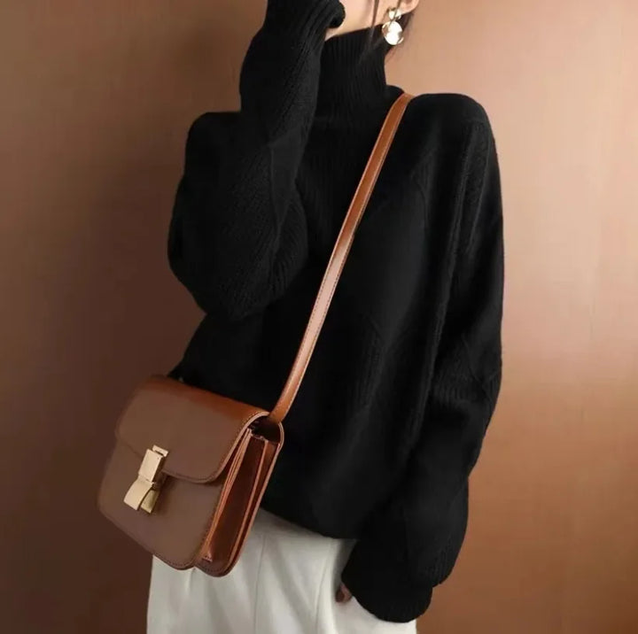 A person wearing a black cashmere turtleneck sweater, white pants, and large round earrings is carrying a brown leather shoulder bag with a gold clasp against a plain brown background.