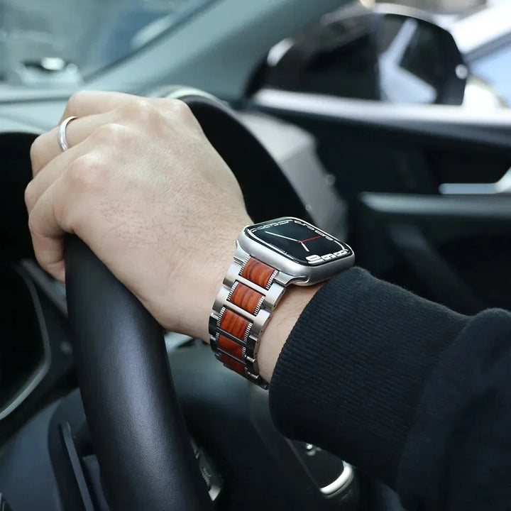 A person wearing a smartwatch with an adjustable Sandalwood watch band grips a steering wheel inside a car. The person is dressed in a black sleeve and wears a silver ring on their finger.