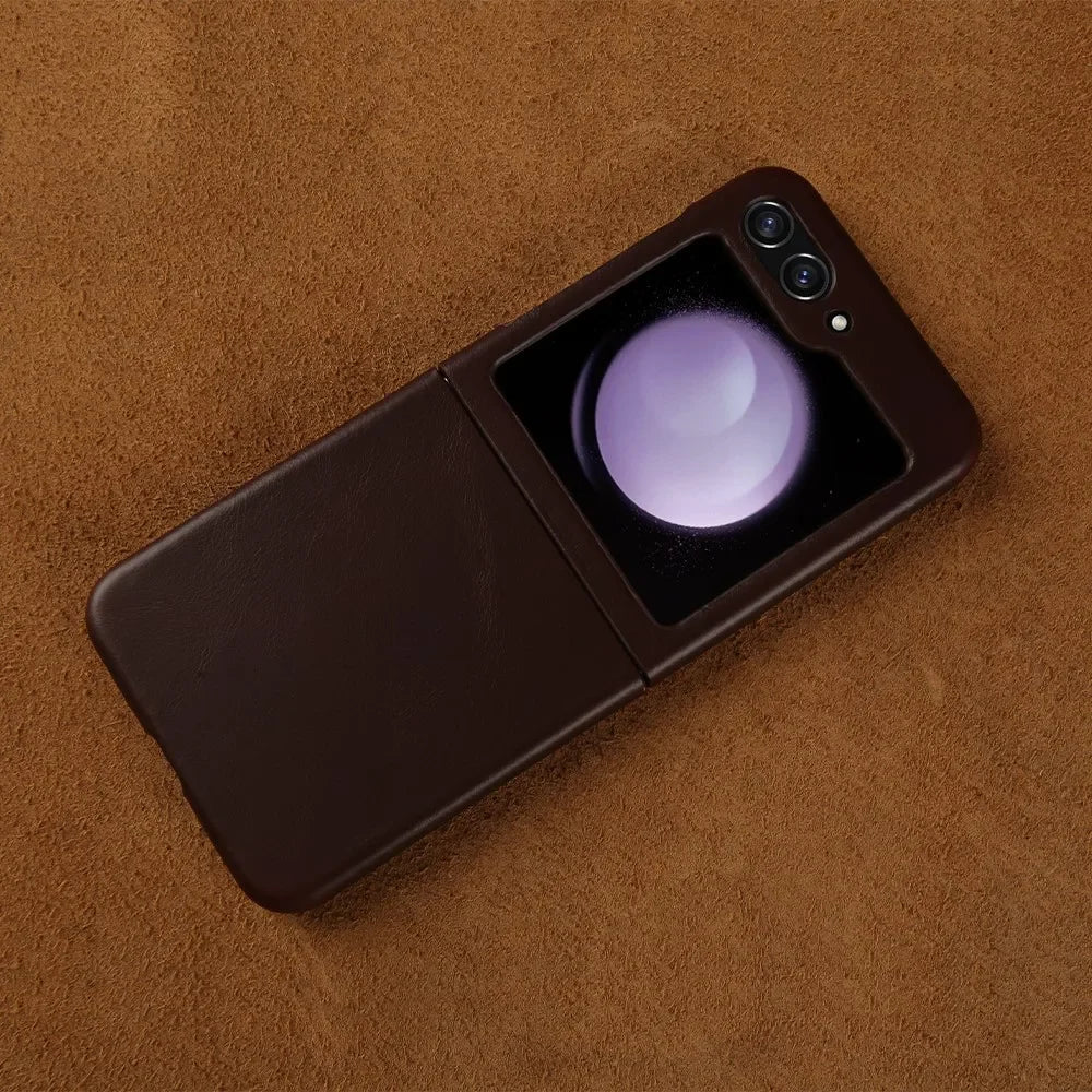 A brown Samsung Galaxy Z Flip foldable smartphone with dual rear cameras and a small external display showing a glowing orb, features genuine leather finish and is placed on a brown textured surface.