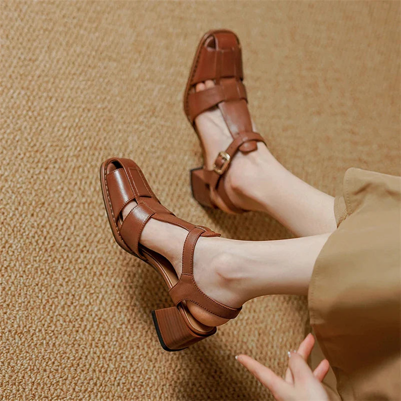 A person wearing handcrafted light brown heeled sandals with ankle straps and wide pants sits on a textured beige carpet, legs crossed at the ankles.