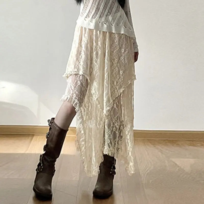 A person wearing a white, long-sleeve, ribbed top paired with a high-waist cream skirt featuring a silky lace asymmetrical hemline and brown mid-calf boots with buckles stands indoors on a wooden floor against a plain light wall.