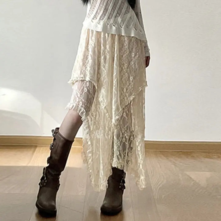 A person wearing a white, long-sleeve, ribbed top paired with a high-waist cream skirt featuring a silky lace asymmetrical hemline and brown mid-calf boots with buckles stands indoors on a wooden floor against a plain light wall.