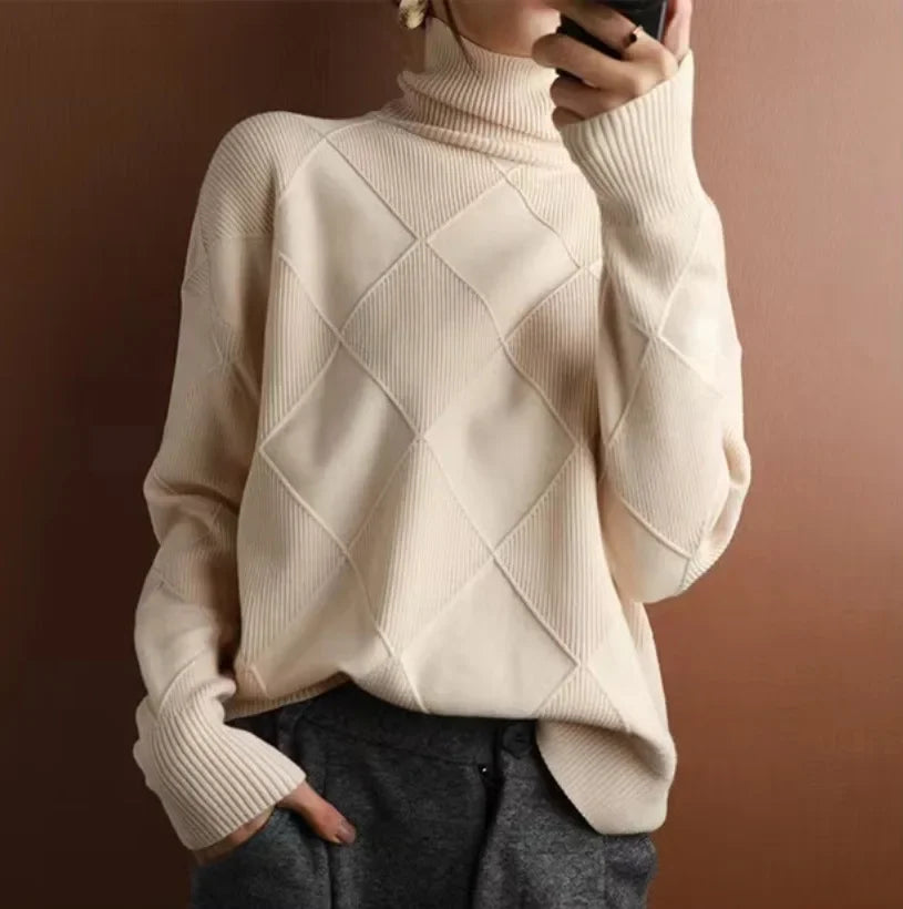 A person wearing a cream-colored, diamond knit turtleneck sweater with a ribbed texture, paired with dark gray pants, stands against a brown background and holds a phone up for a mirror selfie.