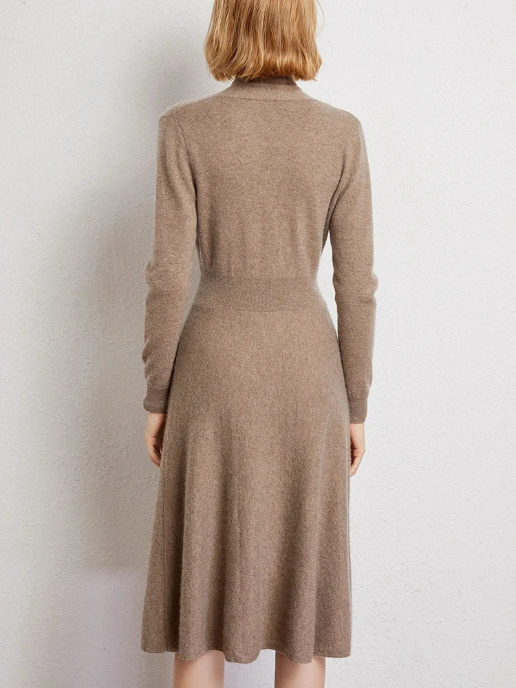A woman with short blonde hair, seen from behind, wears a long-sleeved, knee-length Matilda Wrap Dress in light brown knit and stands against a plain, light-colored wall.