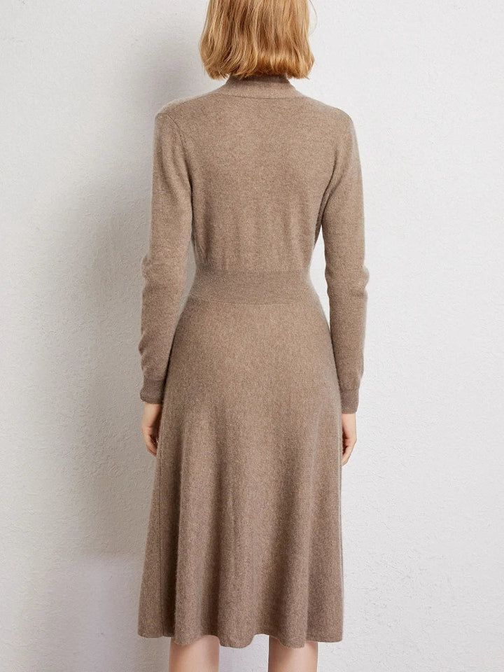 A woman with short blonde hair, seen from behind, wears a long-sleeved, knee-length Matilda Wrap Dress in light brown knit and stands against a plain, light-colored wall.