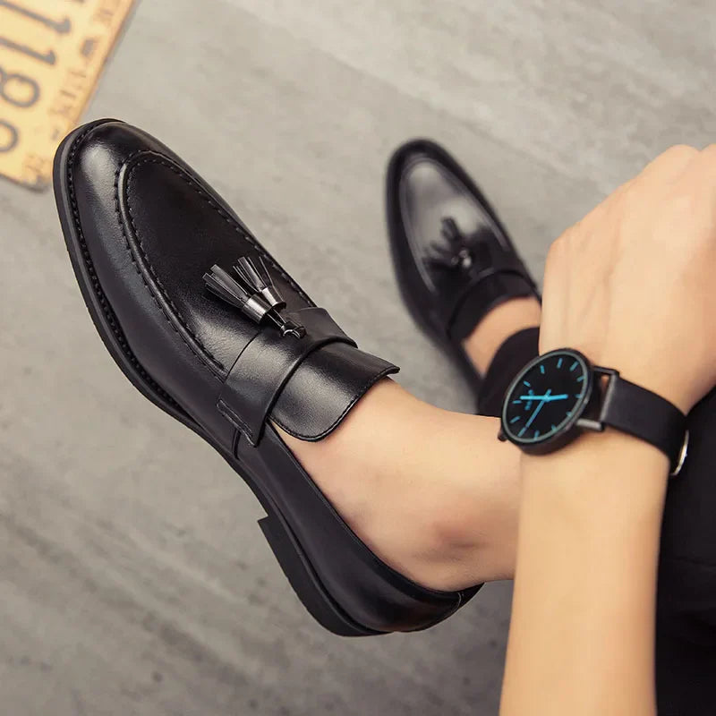 A person wearing minimalistic polished leather tassel loafers and a black wristwatch with a blue face sits on a gray floor, with one leg crossed over the other.