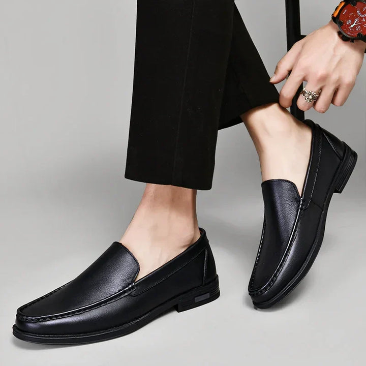 A person wearing black dress pants and classic slip-on genuine leather loafers, sitting with one leg crossed over the other. The plain gray background highlights the sophistication of the shoes.