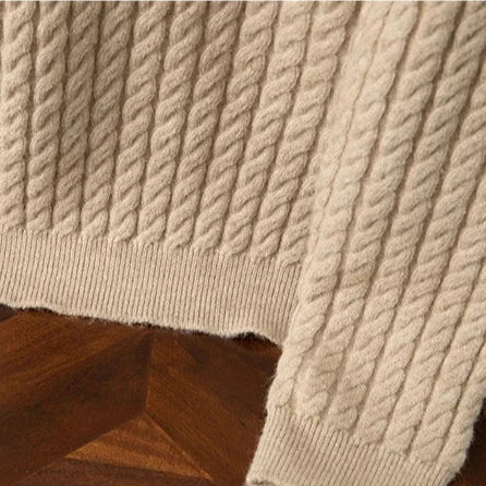Close-up of a beige cable-knit sweater sleeve and hem, showing textured, twisted knit patterns and ribbed cuffs, laid on a wooden surface with a geometric pattern.