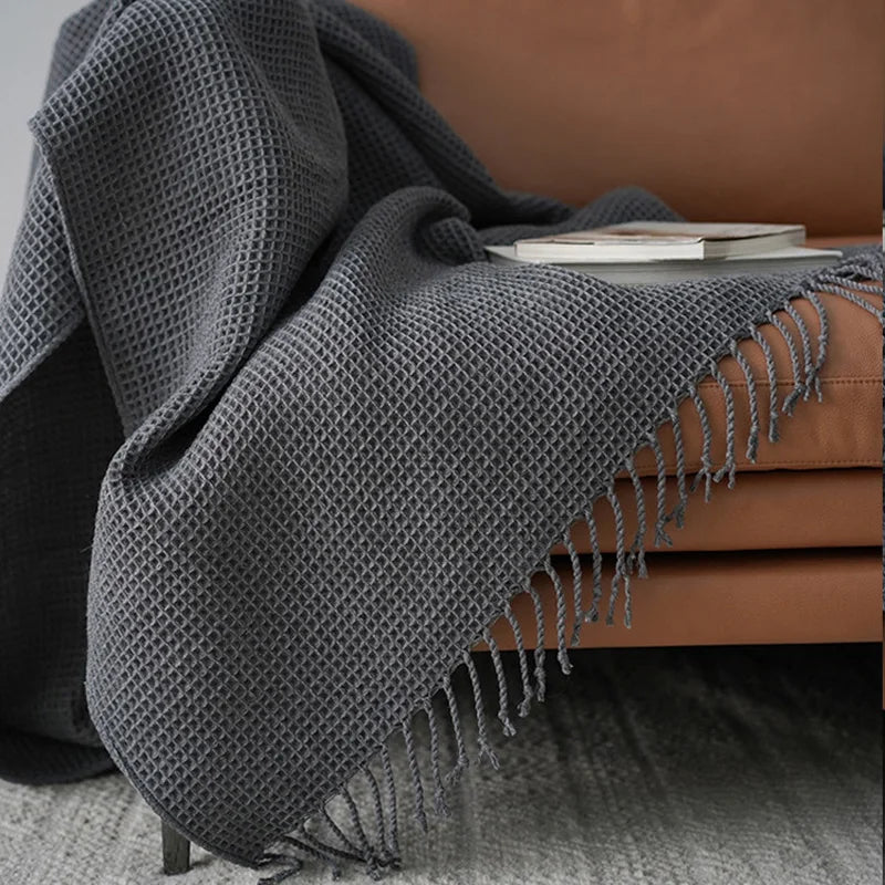 A textured knit waffle blanket with fringe is draped over a brown leather chair beside a closed book, with part of the blanket touching a gray carpeted floor.