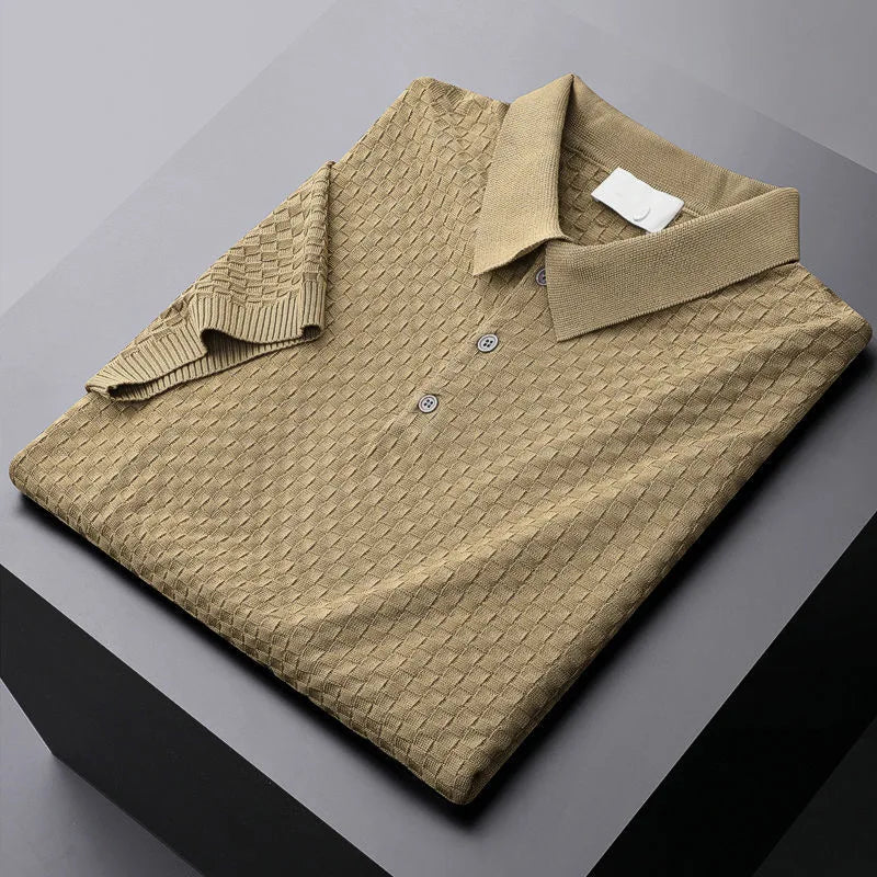 A folded light brown waffle jacquard polo shirt with a collar and three buttons rests on a dark gray surface. One sleeve is laid diagonally across the shirt, highlighting its detailed handmade pattern.