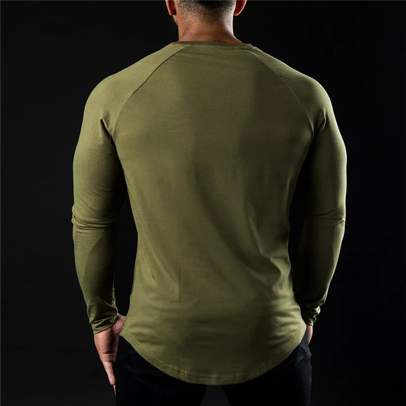 A man is standing with his back to the camera, wearing a long sleeve fitted shirt in olive green and black pants against a plain black background.