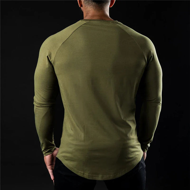 A man is standing with his back to the camera, wearing a long sleeve fitted shirt in olive green and black pants against a plain black background.