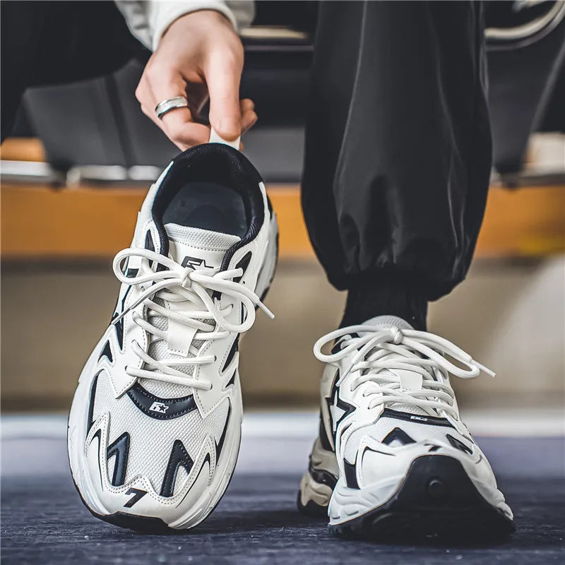 A person wearing black pants and a ring sits down, tying the laces on white and black athletic sneakers with a durable rubber outsole. The focus is on the shoes and hands.