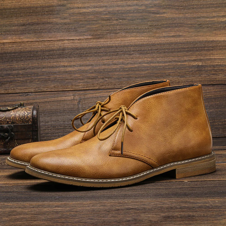 A pair of tan leather After-Ride Chukka boots with laces on a wooden surface, featuring visible stitching along the soles.