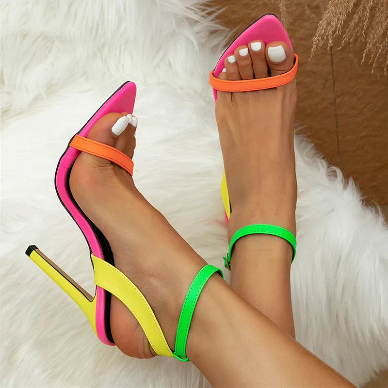 A pair of feet wearing high-heeled sandals with a fashion-forward design, featuring neon pink, orange, yellow, and green straps. The person has white toenail polish and is sitting on a fluffy white surface.