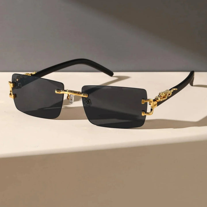 A pair of stylish sunglasses with rectangular black lenses, rimless frames, gold accents on the durable hinges, and black arms, displayed on a white surface against a neutral background.